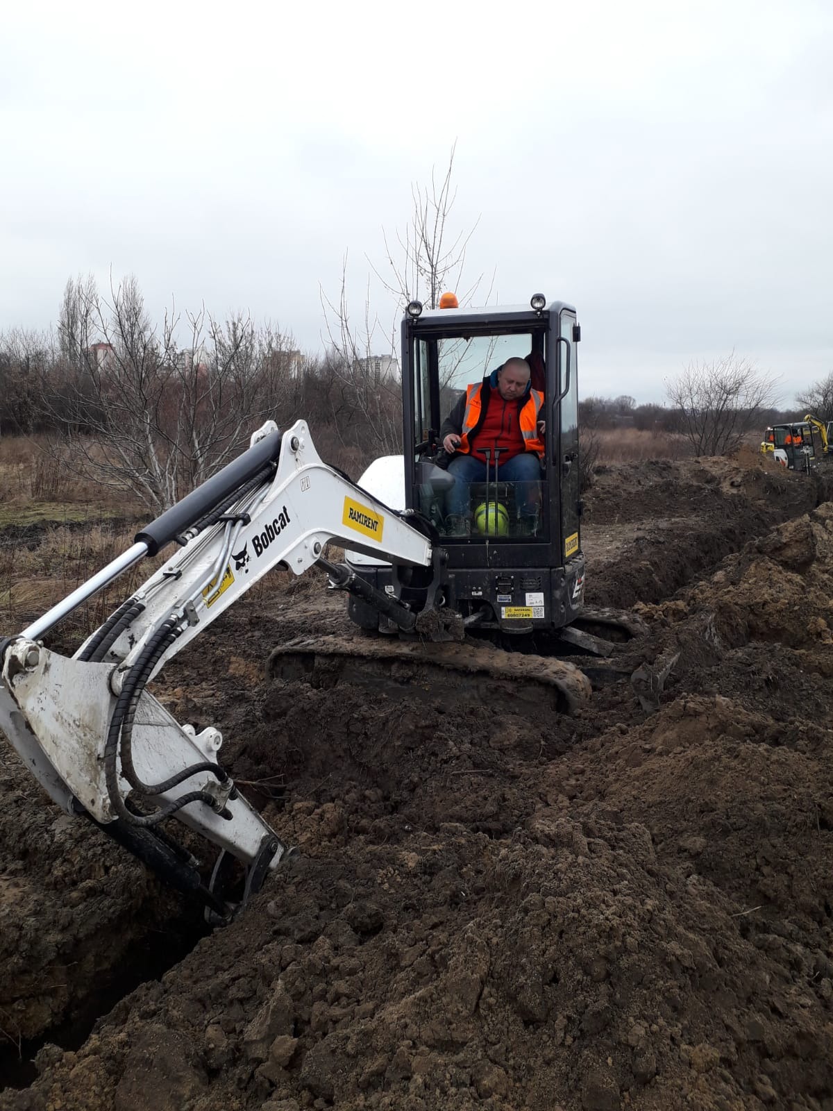Operator w minikoparce Bobcat wykonuje wykop w ziemi, na tle widoczne drzewa bez liści i inne maszyny budowlane. Praca w pochmurny dzień.