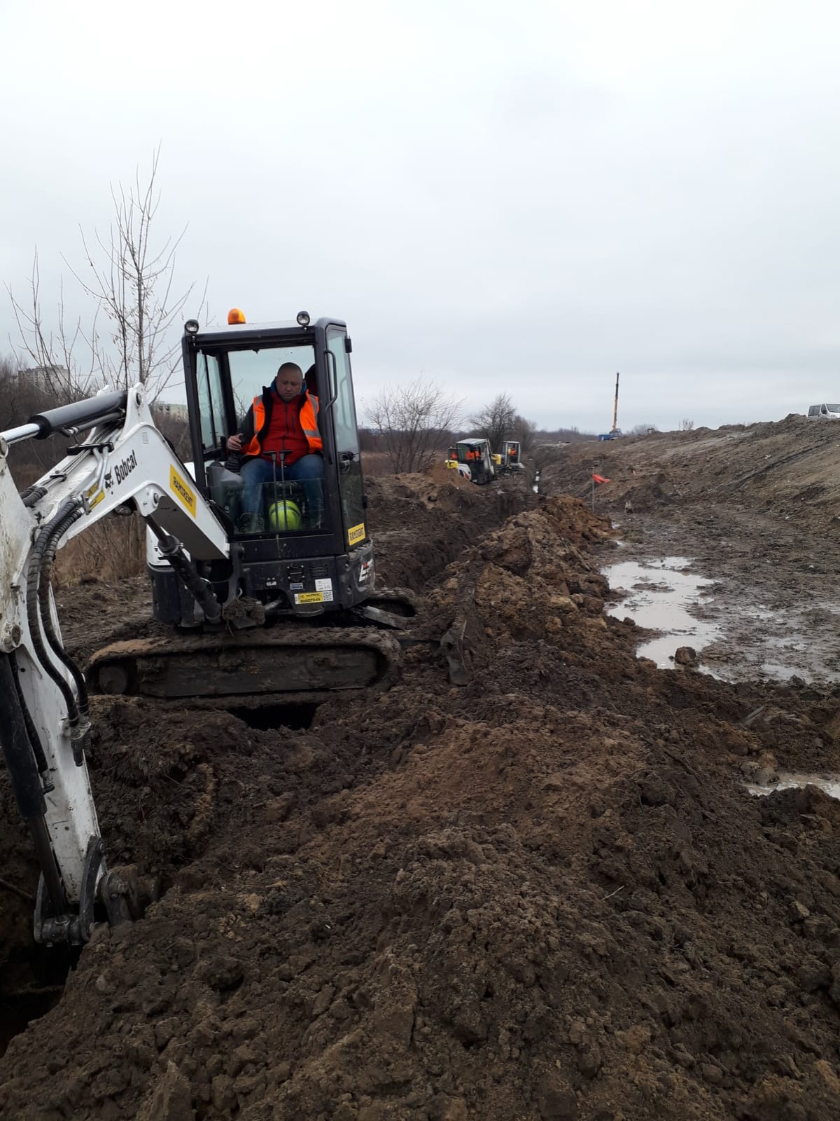 Praca mini koparki Bobcat przy wykopie rowu w grząskim terenie; operator w pomarańczowej kamizelce, w tle inne maszyny i krajobraz.