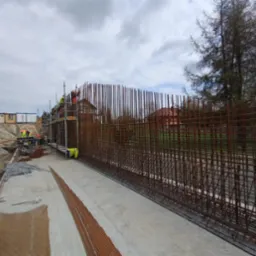 Zbrojenie fundamentów pod budowę, widoczne rusztowania i pracownicy w odblaskowych ubraniach. Zbrojenie wykonane z prętów stalowych, tło: budynki mieszkalne.