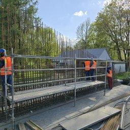 Job Nest sp. z.o.o - Zbrojenie fundamentów pod budowę domu jednorodzinnego. Trzech pracowników w kaskach i kamizelkach na rusztowaniu. Widoczne pręty zbrojeniowe i elementy konstrukcyjne.