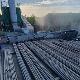 Job Nest sp. z.o.o - Przygotowane zbrojenie na placu budowy, wiązki prętów i gotowe szkielety zbrojeniowe, w tle budynek przemysłowy z kominem i lekka mgła.