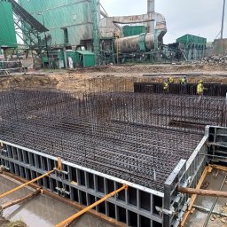 Job Nest sp. z.o.o - Zbrojenie fundamentów pod budowę przemysłową z widoczną konstrukcją szalunkową i pracownikami w oddali, na tle zielonej infrastruktury przemysłowej.