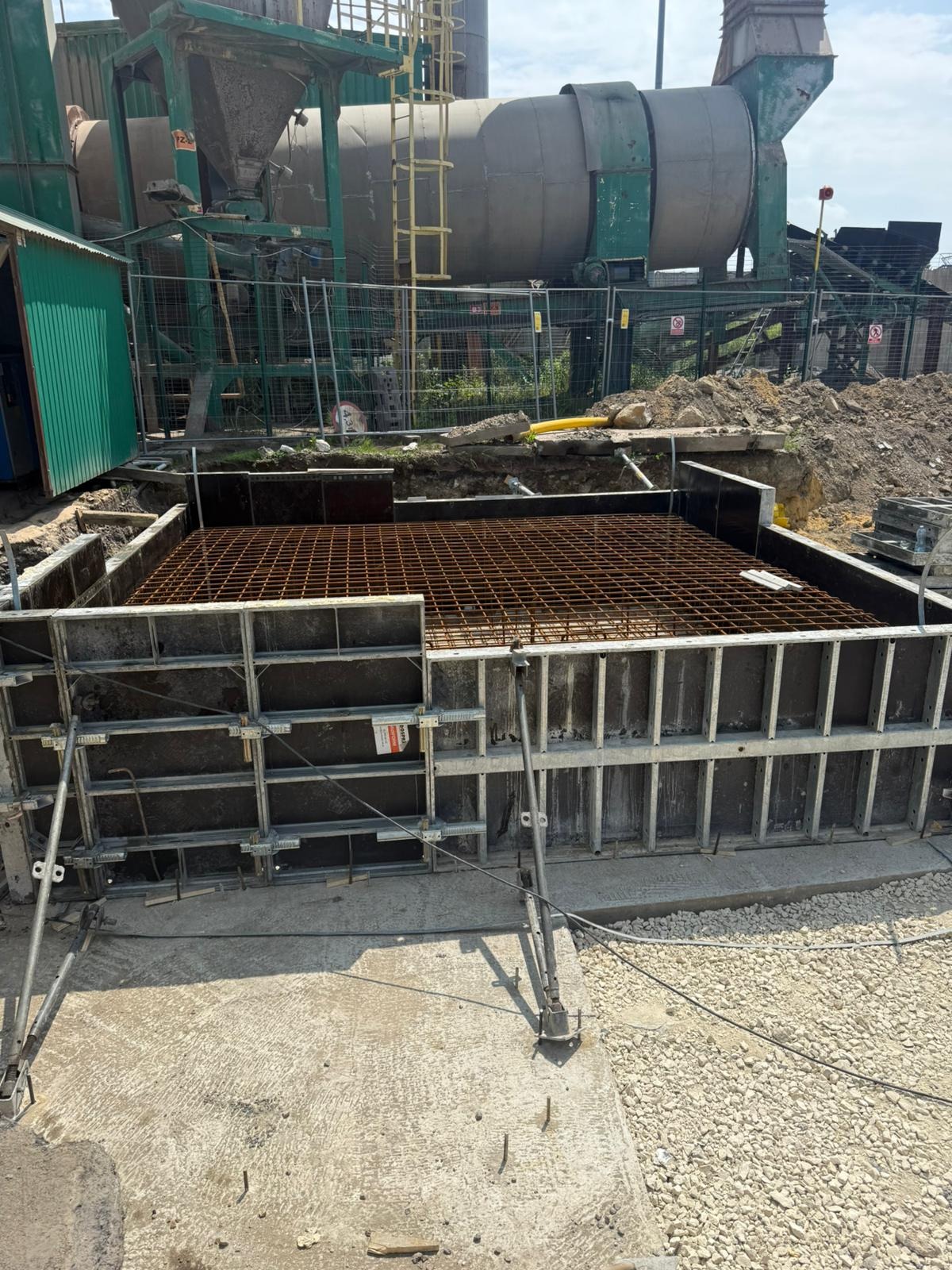 Szare szalunki fundamentowe z ułożonym zbrojeniem na placu budowy. W tle betoniarka i zielony barak.