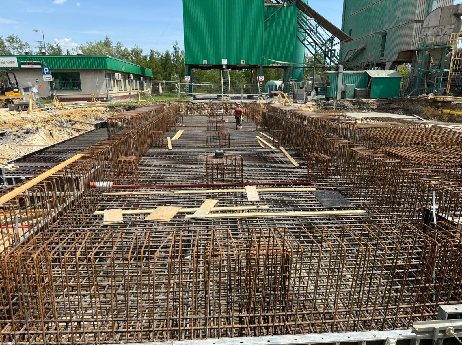 Siatka zbrojeniowa w wykopie pod fundamenty, widok z góry. Na drugim planie budynek przemysłowy z zielonym dachem. Prace ziemne w toku, widoczny człowiek.