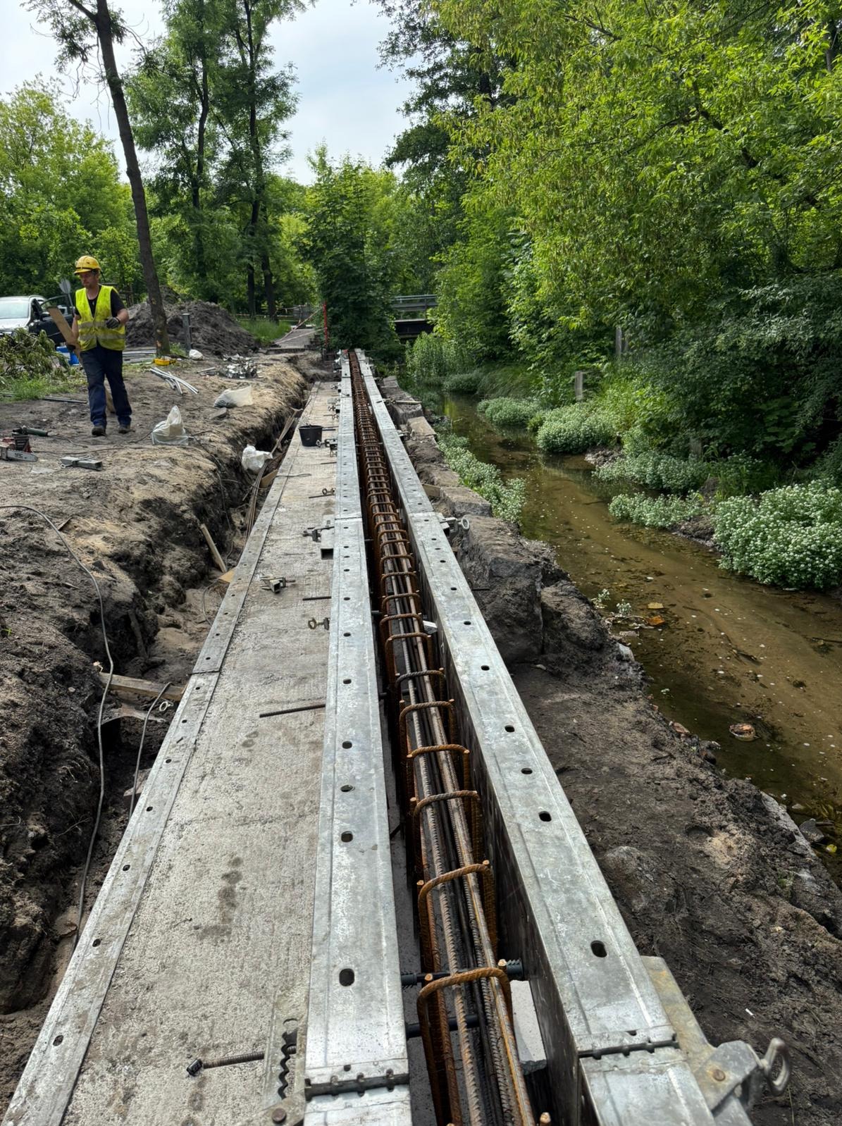 Długa forma zbrojeniowa z widoczną stalą zbrojeniową, przygotowana do zalania betonem. W tle rzeka i pracownik w kasku. Widok z góry.