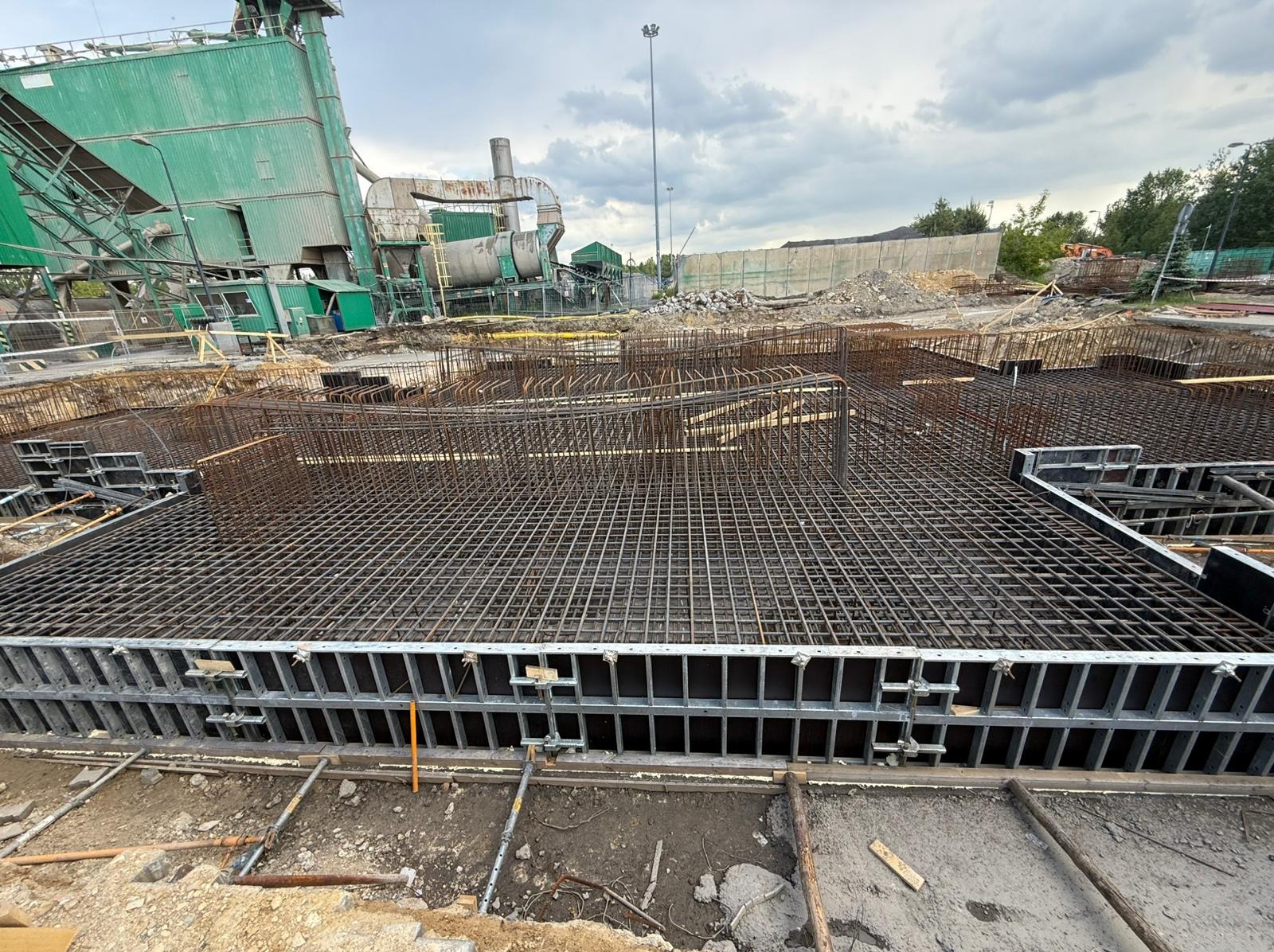Zbrojenie fundamentów z szalunkiem metalowym, widok z góry na plac budowy z zielonym budynkiem przemysłowym w tle.