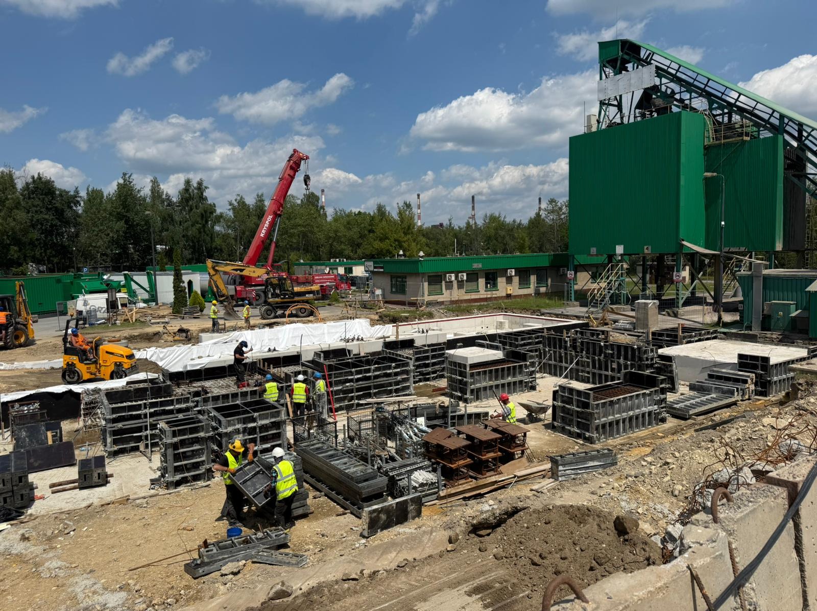 Budowa fundamentów: pracownicy w kaskach montują szalunki pod żelbet, widoczny dźwig i maszyny budowlane na tle zielonej hali i błękitnego nieba.