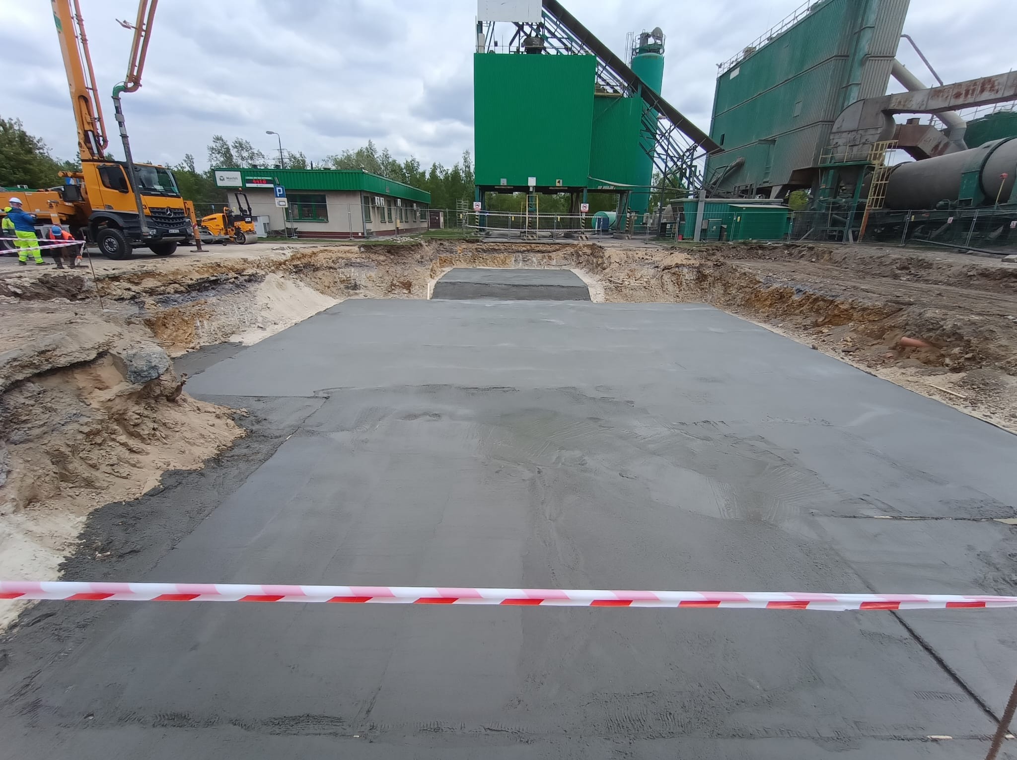 Wylany fundament z betonu na budowie, otoczony wykopem i taśmą ostrzegawczą. W tle maszyny budowlane i budynek. Pochmurne niebo.