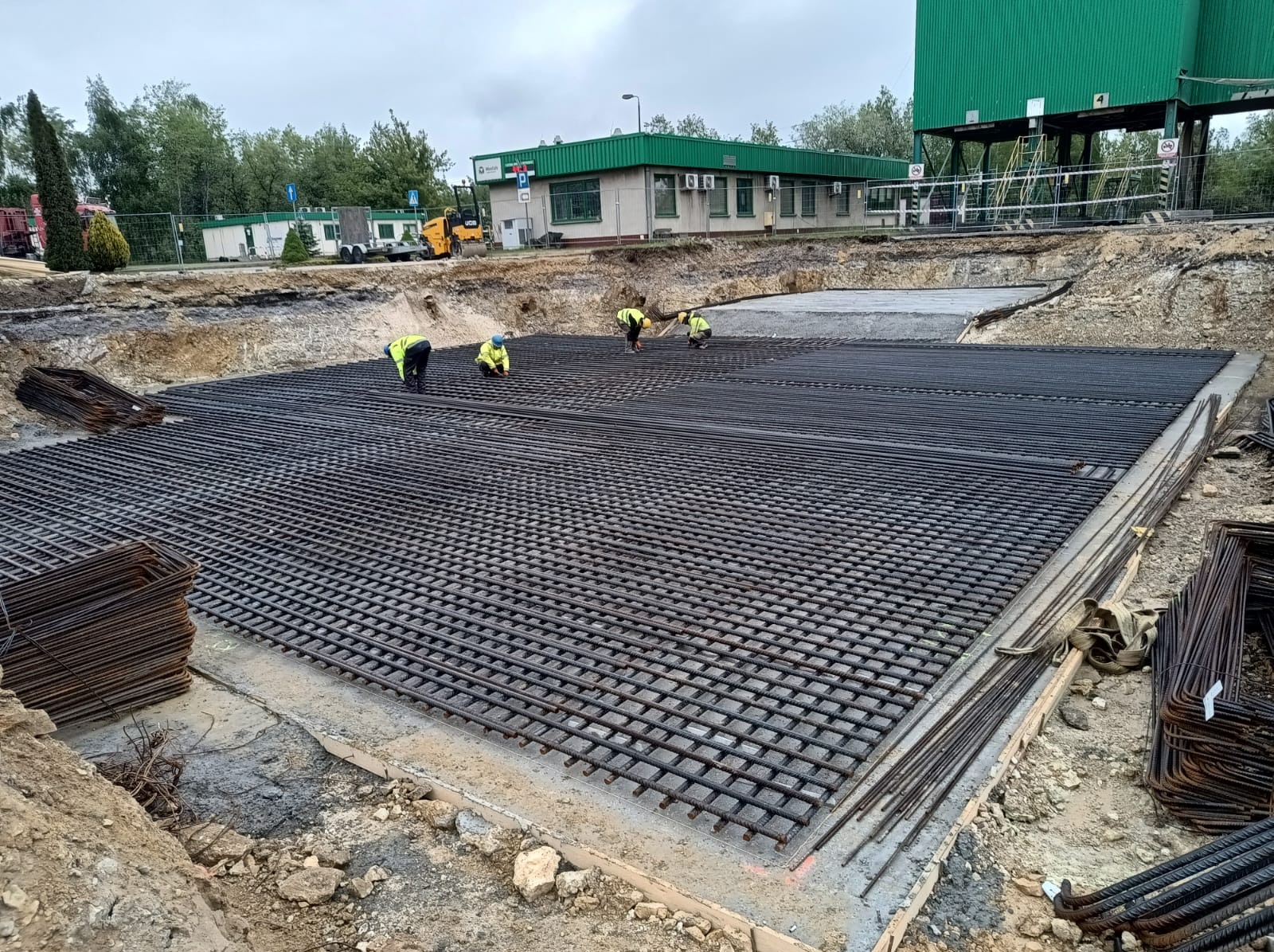 Zbrojenie fundamentu pod budynek przemysłowy. Siatka ze stalowych prętów na betonowej wylewce, robotnicy w żółtych kamizelkach i maszyny budowlane w tle.