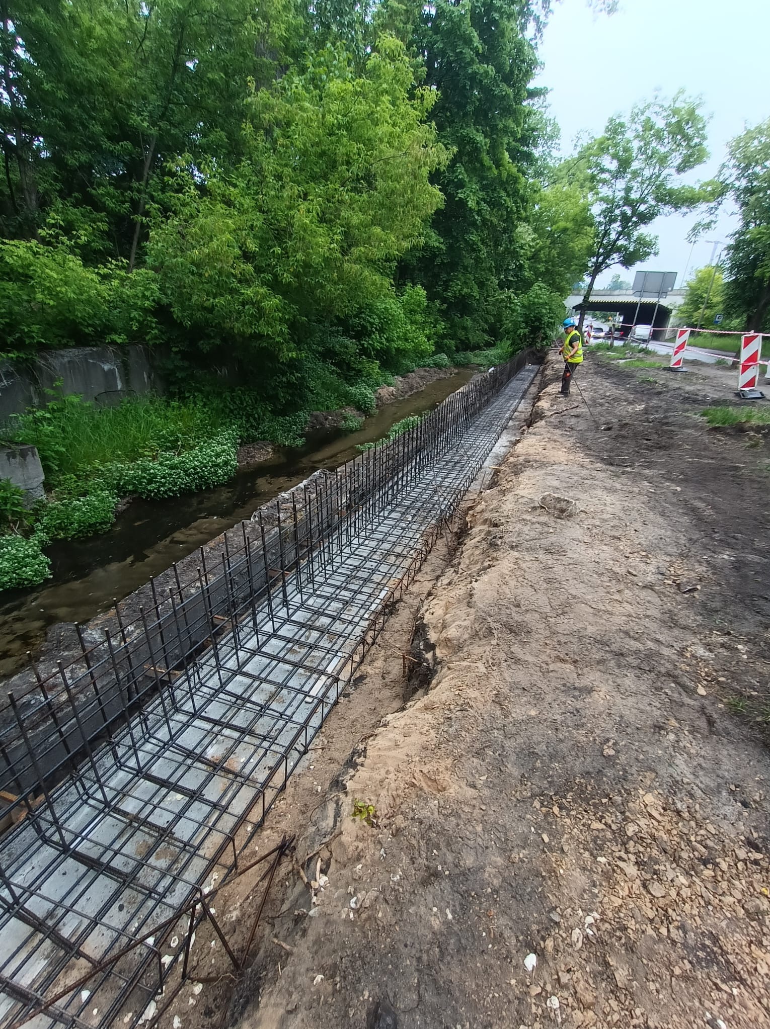 Zbrojenie koryta rzeki z pracownikiem w kamizelce odblaskowej w tle, widok z góry. Prace ziemne obok koryta rzeki.