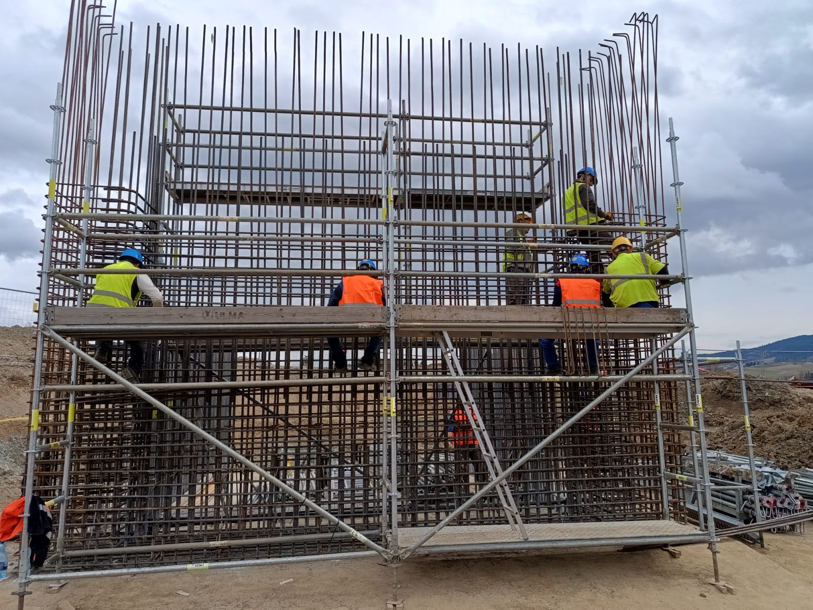 Zbrojenie żelbetowe na budowie w Krakowie. Robotnicy w kaskach i kamizelkach na rusztowaniu, przygotowują konstrukcję z prętów stalowych. Widoczne elementy krajobrazu.