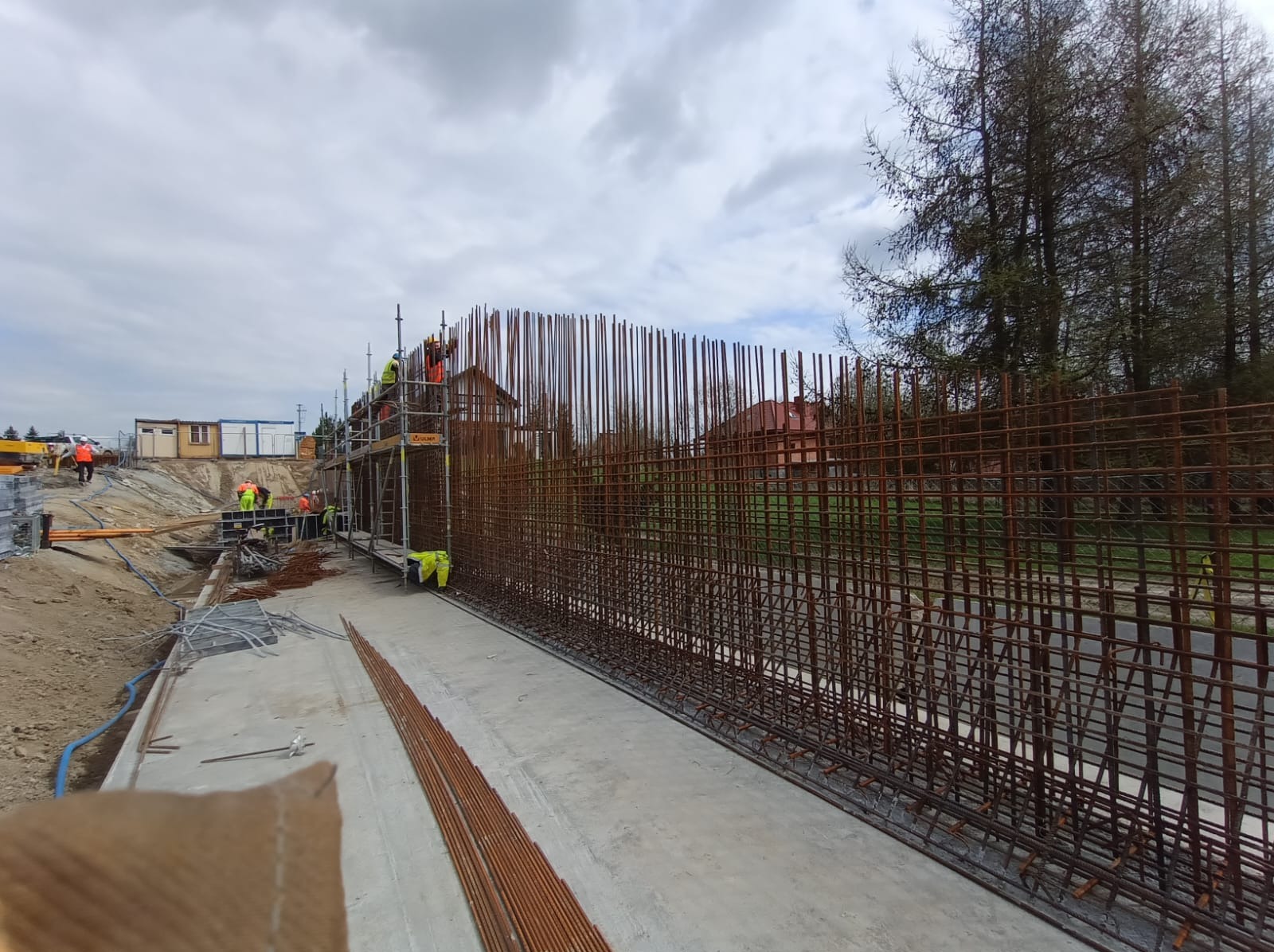 Zbrojenie fundamentów pod budowę, widoczne rusztowania i pracownicy w odblaskowych ubraniach. Zbrojenie wykonane z prętów stalowych, tło: budynki mieszkalne.
