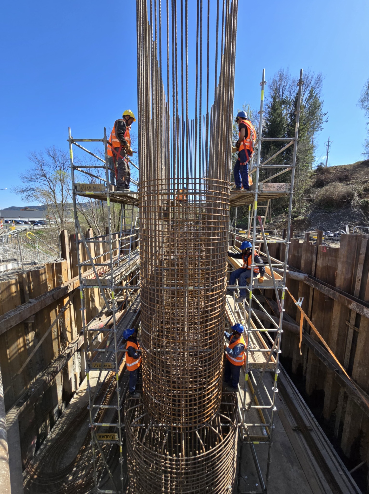 Montaż zbrojenia kolumny na budowie. Robotnicy w kaskach i kamizelkach na rusztowaniach, widok z dołu na konstrukcję z prętów stalowych.