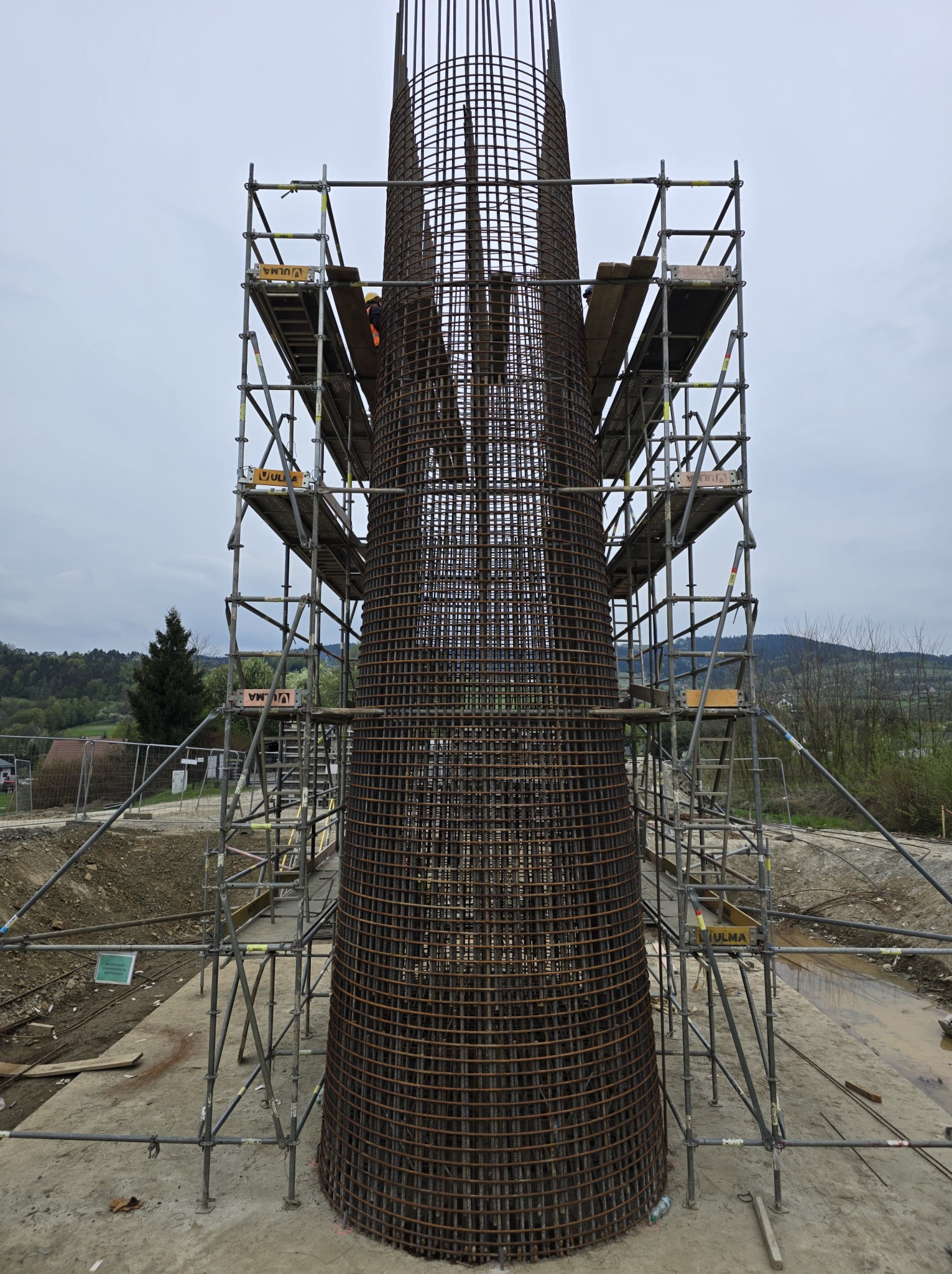 Zbrojenie kolumny na budowie, otoczone rusztowaniem. Widoczny pracownik w kasku i kamizelce. Konstrukcja na tle zieleni i pochmurnego nieba.