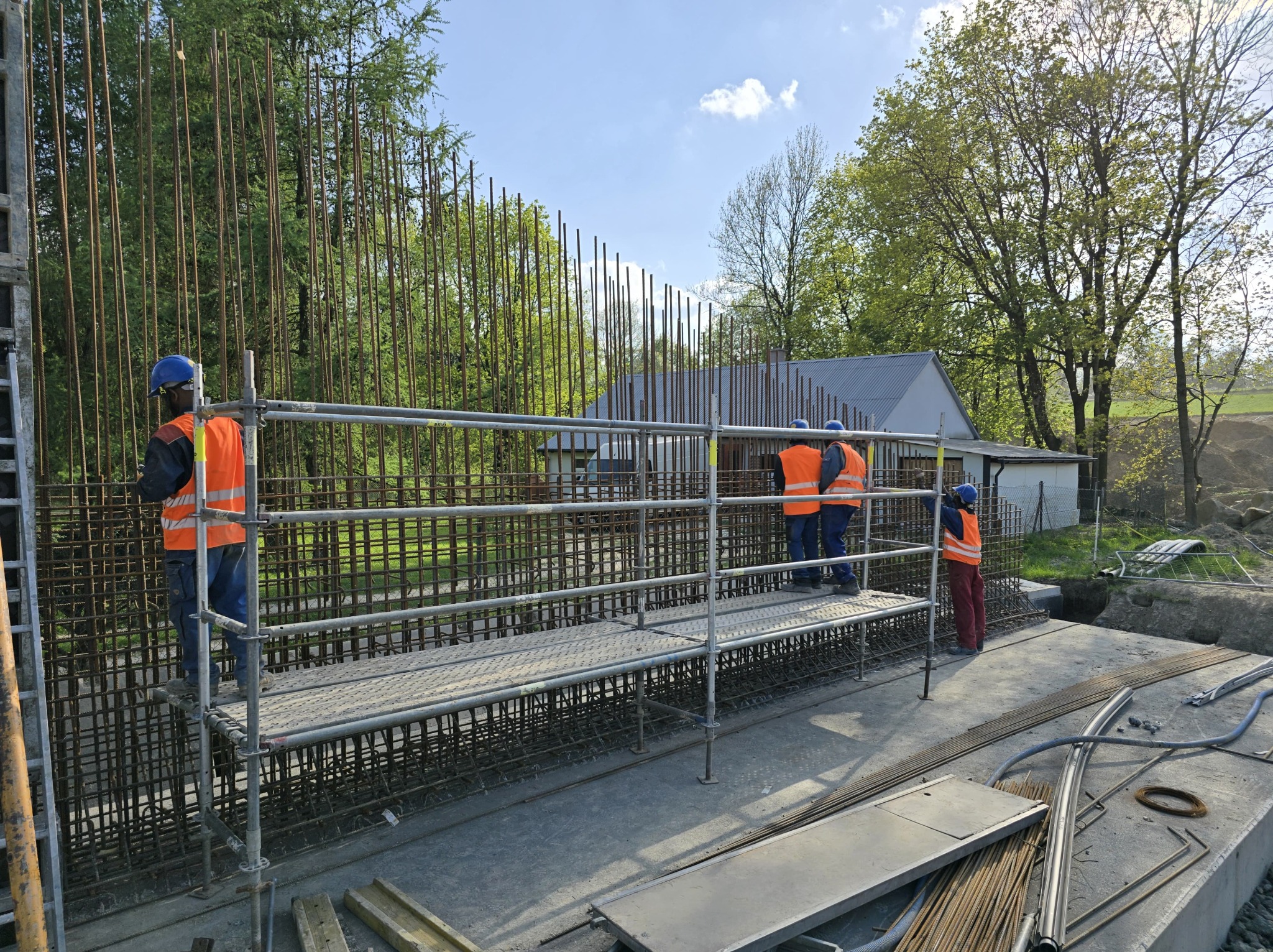 Zbrojenie fundamentów pod budowę domu jednorodzinnego. Trzech pracowników w kaskach i kamizelkach na rusztowaniu. Widoczne pręty zbrojeniowe i elementy konstrukcyjne.