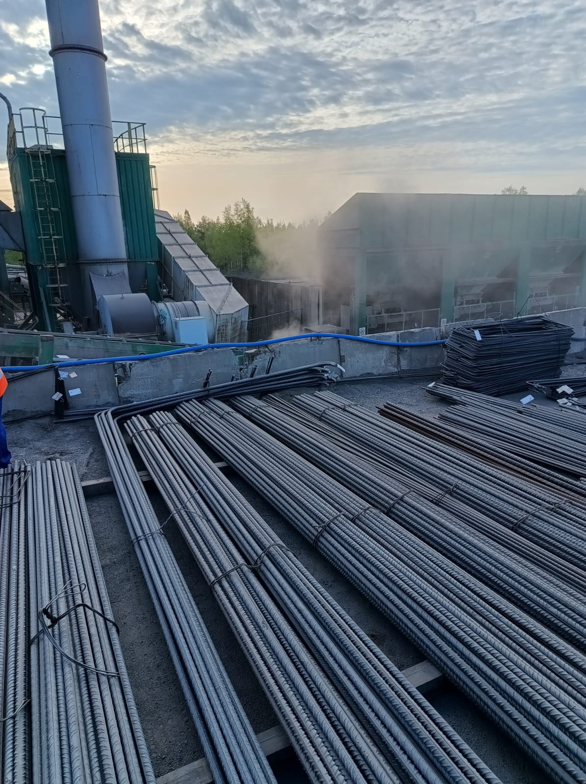 Przygotowane zbrojenie na placu budowy, wiązki prętów i gotowe szkielety zbrojeniowe, w tle budynek przemysłowy z kominem i lekka mgła.