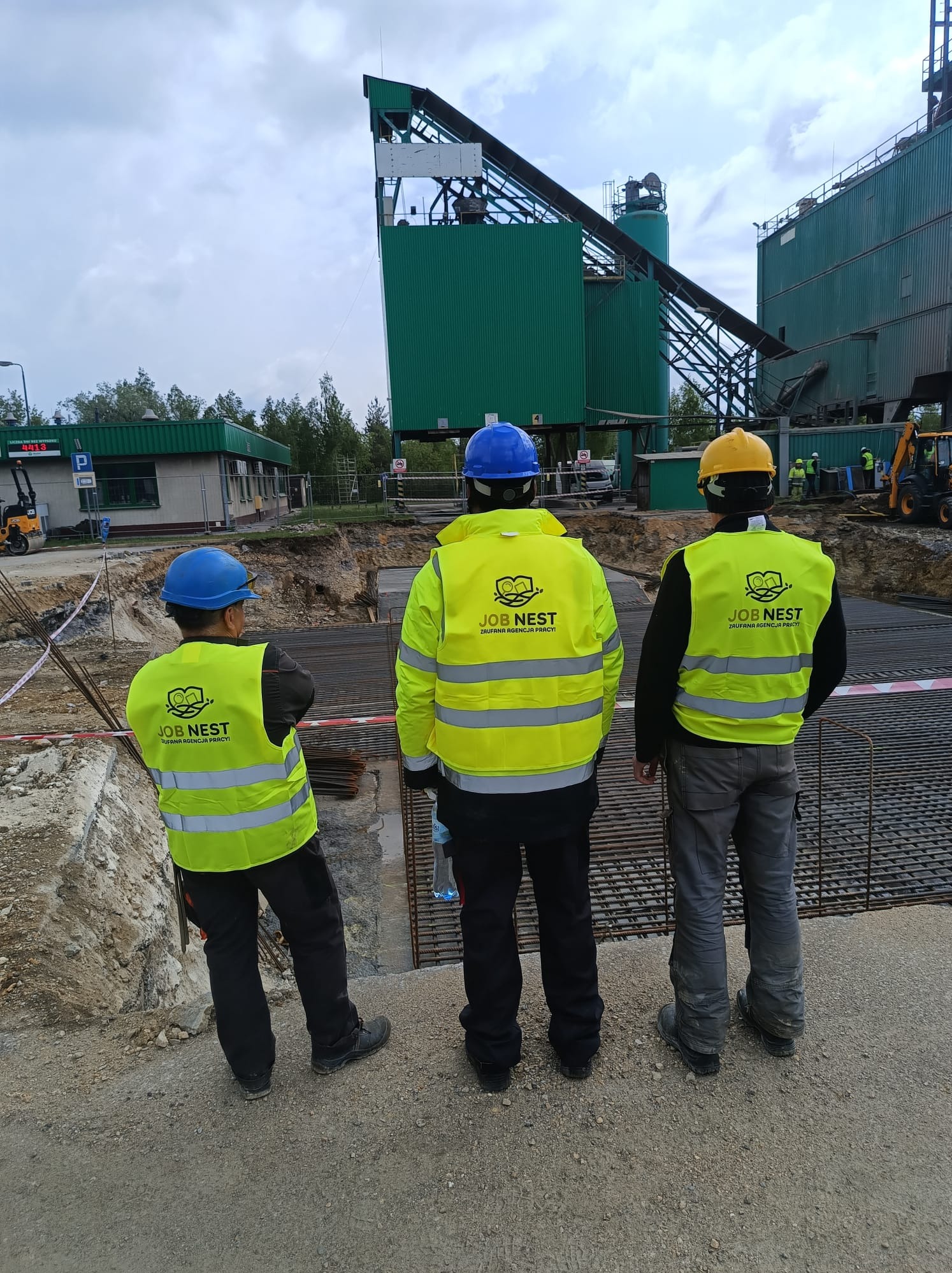 Pracownicy w odblaskowych kamizelkach obserwują zbrojenie fundamentów pod budynek przemysłowy. Widoczna siatka zbrojeniowa, wykop i maszyny budowlane w tle.