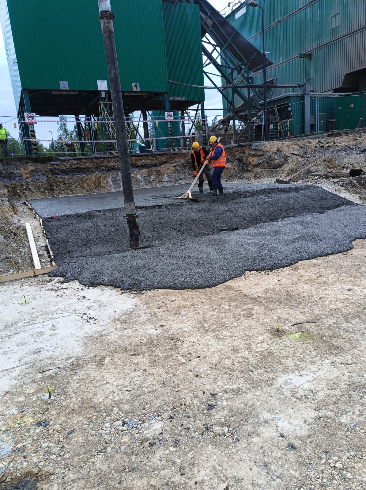 Wylewanie betonu na budowie przemysłowej. Dwóch robotników w kaskach i kamizelkach rozprowadza beton za pomocą drewnianej łaty. W tle zielony budynek przemysłowy.