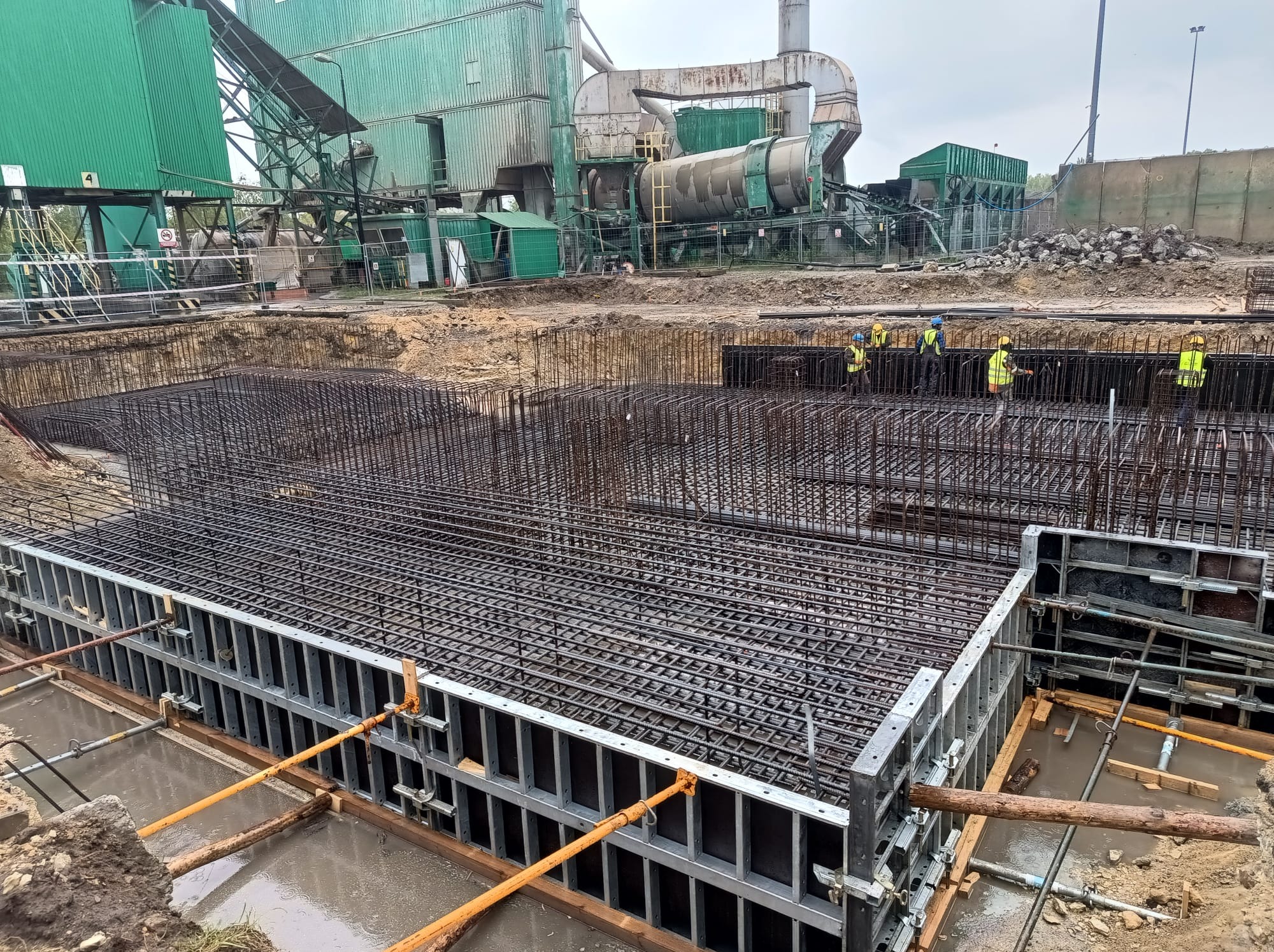 Zbrojenie fundamentów pod budowę przemysłową z widoczną konstrukcją szalunkową i pracownikami w oddali, na tle zielonej infrastruktury przemysłowej.