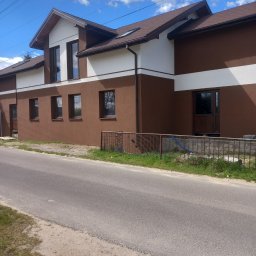 KROC-BUD - Dwukondygnacyjny dom z brązową elewacją i białymi pasami, widoczny z poziomu ulicy w słoneczny dzień. Brązowe okna i dachówka. Metalowe ogrodzenie przed budynkiem.