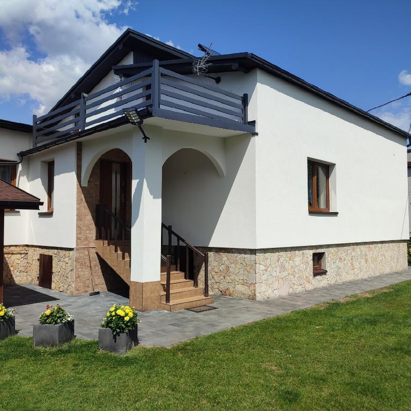 Biała elewacja domu z balkonem i kamienną podmurówką, widok z zewnątrz na tle błękitnego nieba. Eleganckie wykończenie z dbałością o detale.