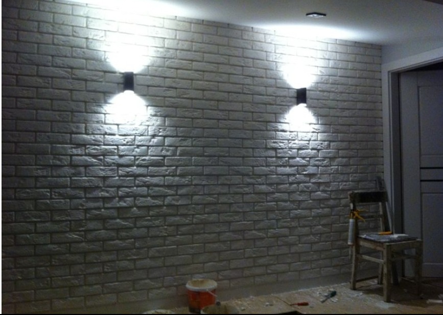 Ściana z dekoracyjnej cegły w jasnym odcieniu, oświetlona dwoma kinkietami. Widoczny fragment podłogi z drobnymi zabrudzeniami po pracy i krzesło.