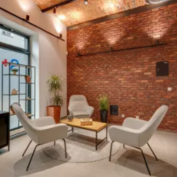 Klimatyczne wnętrze poczekalni z ceglaną ścianą, trzema fotelami i stolikiem kawowym na okrągłym dywanie. Oświetlenie punktowe podkreśla teksturę cegieł.