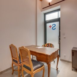 CENTRUM MEDYCZNO-TERAPEUTYCZNE STAMINA SPÓŁKA Z OGRANICZONĄ ODPOWIEDZIALNOŚCIĄ - Gabinet z ceglanym sklepieniem, drewnianym biurkiem i krzesłami w stylu vintage. Na drzwiach logo z plusem i cyfrą 2. Stonowana kolorystyka, minimalistyczny wystrój.