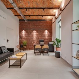 CENTRUM MEDYCZNO-TERAPEUTYCZNE STAMINA SPÓŁKA Z OGRANICZONĄ ODPOWIEDZIALNOŚCIĄ - Nowoczesne wnętrze gabinetu z ceglaną ścianą, minimalistycznym biurkiem, czarną sofą i regałem z dekoracjami roślinnymi. Jasne płytki podłogowe, sufit z widoczną instalacją.