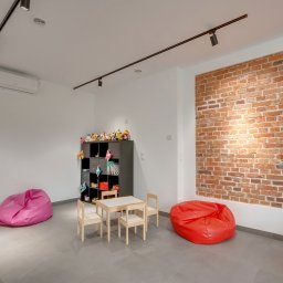 CENTRUM MEDYCZNO-TERAPEUTYCZNE STAMINA SPÓŁKA Z OGRANICZONĄ ODPOWIEDZIALNOŚCIĄ - Jasne wnętrze bawialni z ceglaną ścianą, stolikiem, krzesłami, pufami i regałem z zabawkami. Minimalistyczny design, oświetlenie punktowe, szara podłoga.