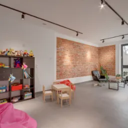 Jasne wnętrze poczekalni z ceglaną ścianą, regałem z zabawkami i stolikiem dla dzieci. W oknie widoczne logo placówki medycznej.