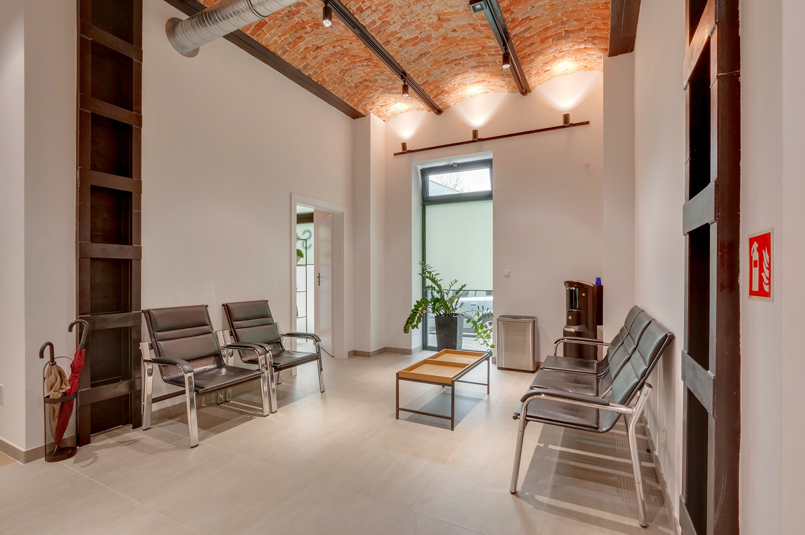 Jasna poczekalnia z ceglanym sklepieniem łukowym, czarnymi krzesłami na metalowych nogach, stolikiem kawowym i rośliną doniczkową przy oknie. Widoczny hydrant.