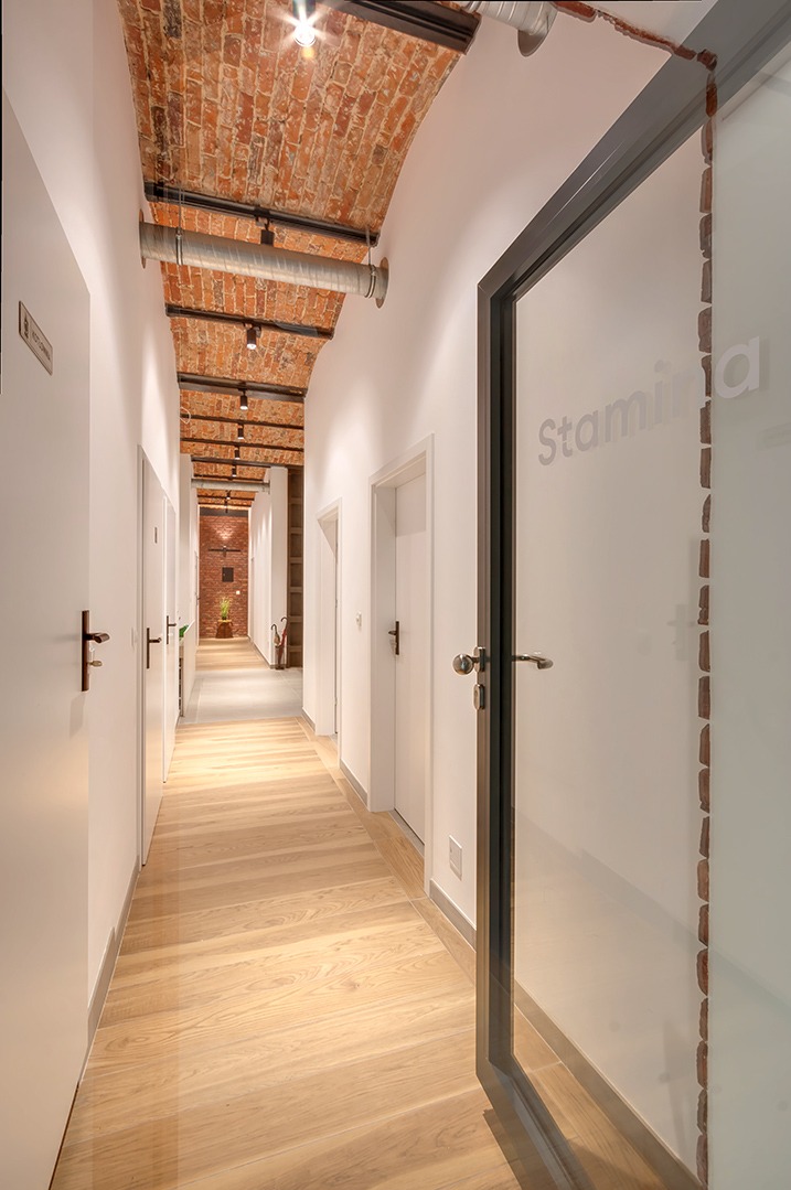 Korytarz w nowoczesnym gabinecie z ceglanym sklepieniem i drewnianą podłogą. Na szklanych drzwiach widnieje napis 'Stamina'. W głębi widoczny jest kwiat w doniczce.