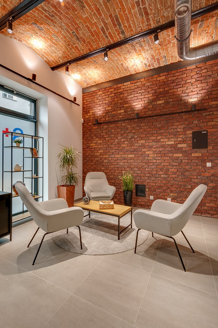 Klimatyczne wnętrze poczekalni z ceglaną ścianą, trzema fotelami i stolikiem kawowym na okrągłym dywanie. Oświetlenie punktowe podkreśla teksturę cegieł.
