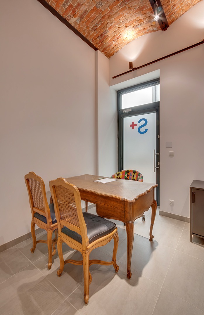 Gabinet z ceglanym sklepieniem, drewnianym biurkiem i krzesłami w stylu vintage. Na drzwiach logo z plusem i cyfrą 2. Stonowana kolorystyka, minimalistyczny wystrój.