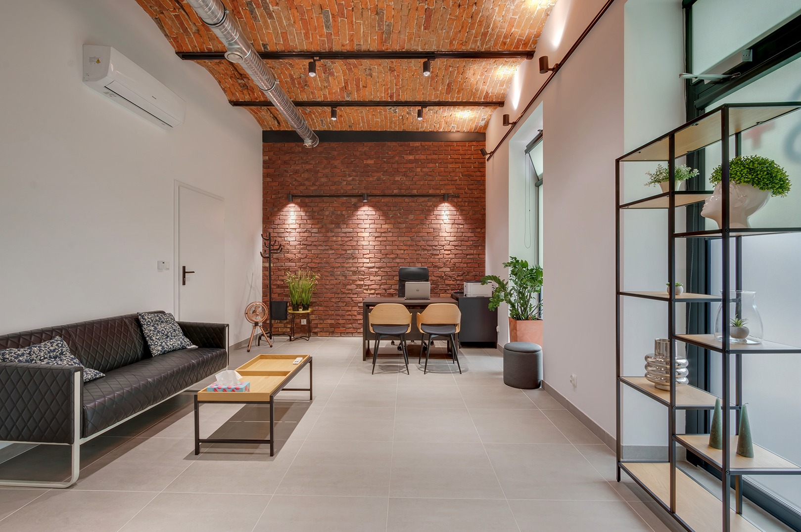 Nowoczesne wnętrze gabinetu z ceglaną ścianą, minimalistycznym biurkiem, czarną sofą i regałem z dekoracjami roślinnymi. Jasne płytki podłogowe, sufit z widoczną instalacją.