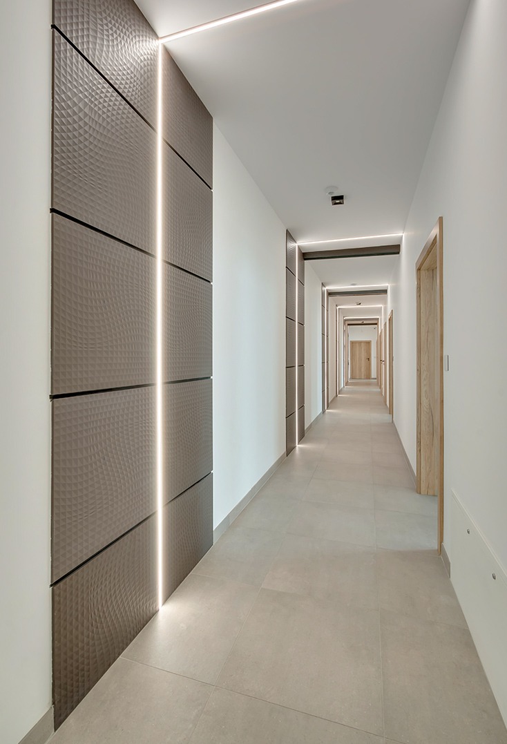 Długi korytarz z panelami ściennymi w odcieniach brązu i oświetleniem LED, płytki podłogowe w kolorze beżowym. Widok w perspektywie, drzwi po prawej stronie.