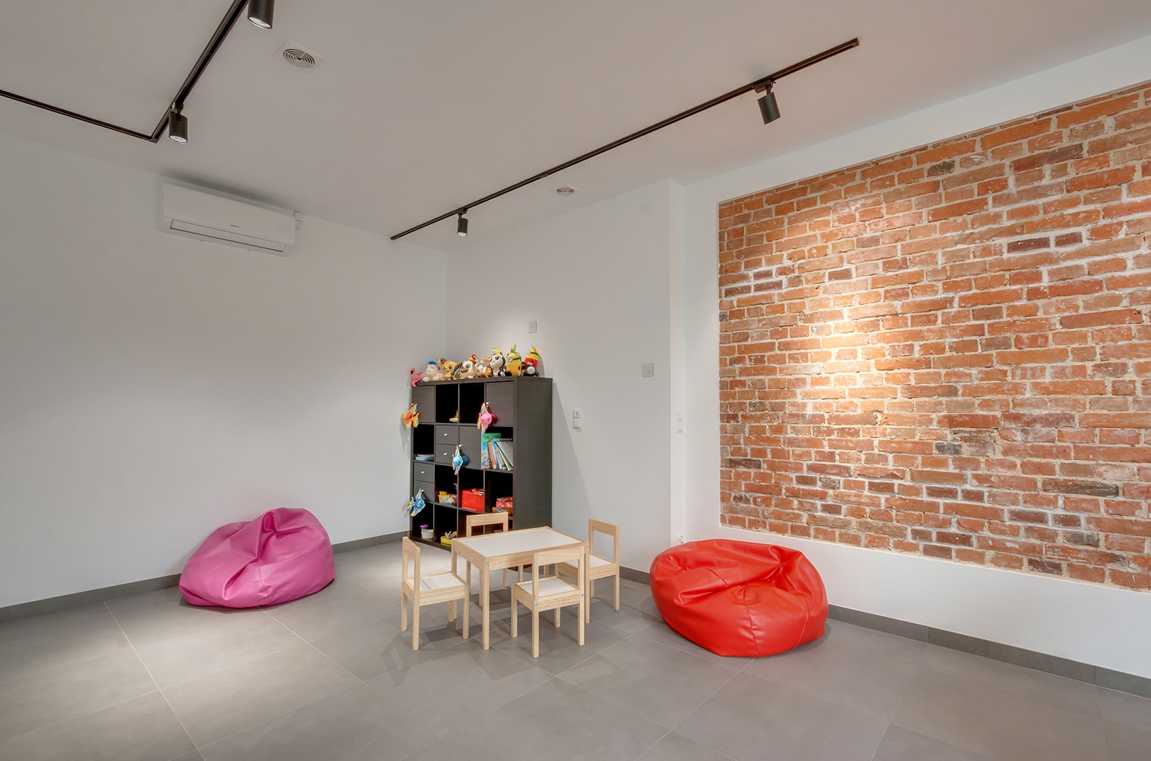 Jasne wnętrze bawialni z ceglaną ścianą, stolikiem, krzesłami, pufami i regałem z zabawkami. Minimalistyczny design, oświetlenie punktowe, szara podłoga.