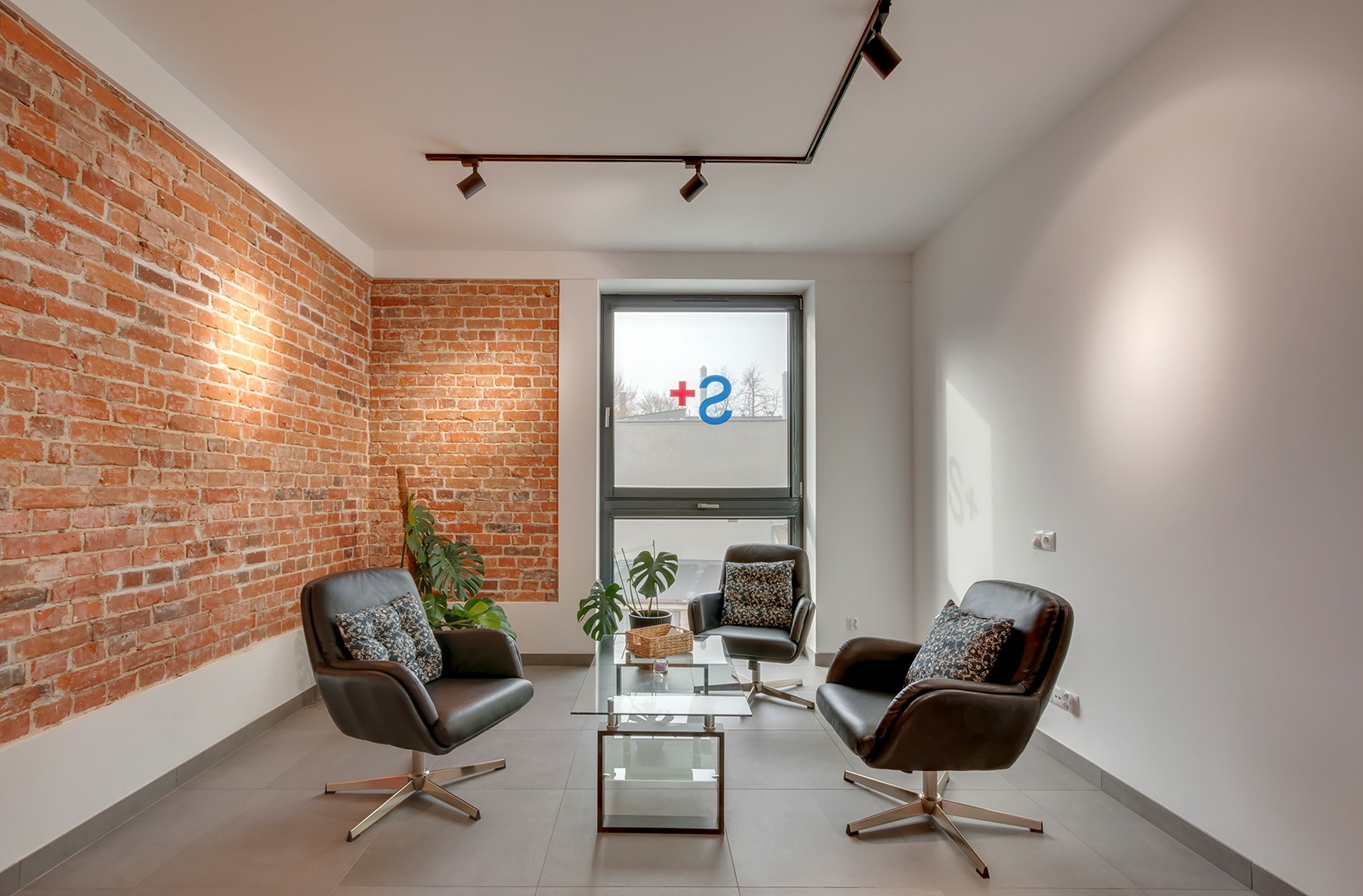 Klimatyczne wnętrze poczekalni z ceglaną ścianą, trzema fotelami i szklanym stolikiem. W oknie widoczne logo firmy. Nowoczesne oświetlenie punktowe.