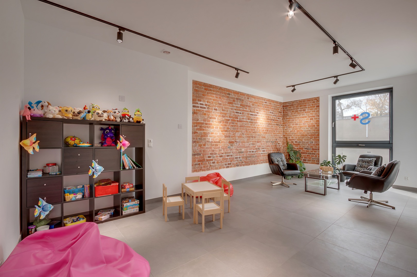 Jasne wnętrze poczekalni z ceglaną ścianą, regałem z zabawkami i stolikiem dla dzieci. W oknie widoczne logo placówki medycznej.