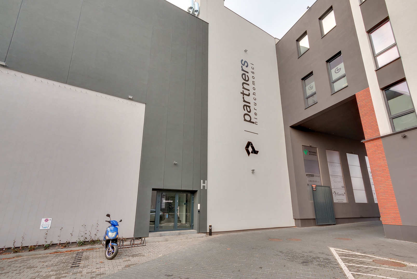 Nowoczesny budynek biurowy z logo 'partners nieruchomości' na fasadzie, skuter zaparkowany obok wejścia. Minimalistyczna architektura w odcieniach szarości i bieli.