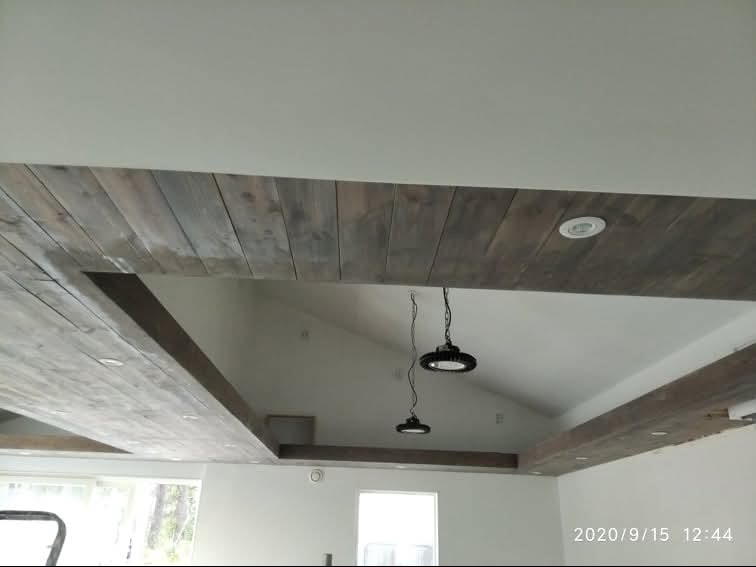 Wykończony sufit z drewnianymi belkami w szarym odcieniu, z wbudowanymi punktowymi lampami i wiszącymi lampami przemysłowymi. Białe ściany i skosy sufitowe.