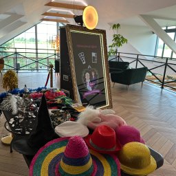 Fotolustro Kuźnia Zdjęć - Fotobudka z lustrem i rekwizytami: kolorowe kapelusze, boa, maski na stole. W tle widoczne eleganckie wnętrze z oknami i roślinami.