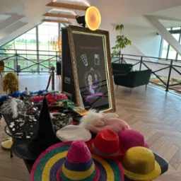 Fotobudka z lustrem i rekwizytami: kolorowe kapelusze, boa, maski na stole. W tle widoczne eleganckie wnętrze z oknami i roślinami.