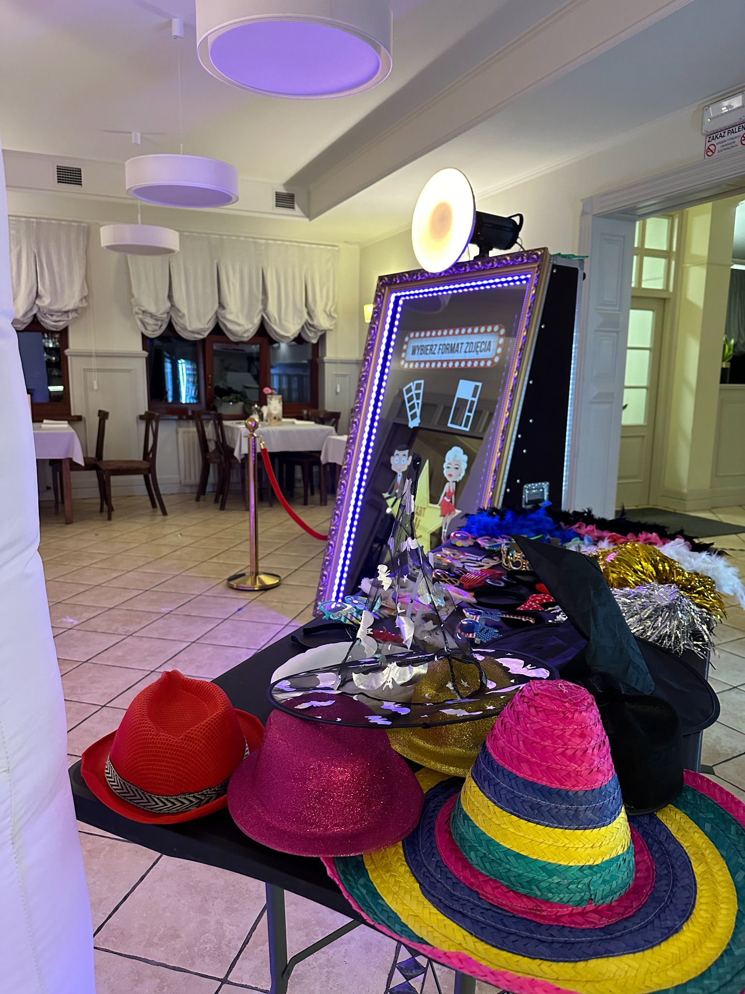 Fotobudka z rekwizytami: kolorowe kapelusze, boa z piór, ramka z napisem 'Wybierz format zdjęcia', tło bankietowe z zasłonami i stolikami.