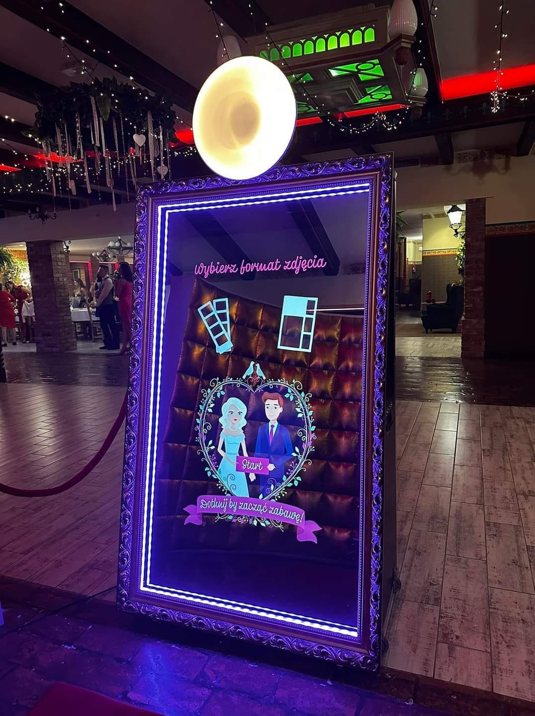 Interaktywna fotobudka w kształcie lustra z animowaną grafiką pary młodej i napisem 'Dotknij by zacząć zabawę!', otoczona dekoracyjną ramą z ledowym oświetleniem.