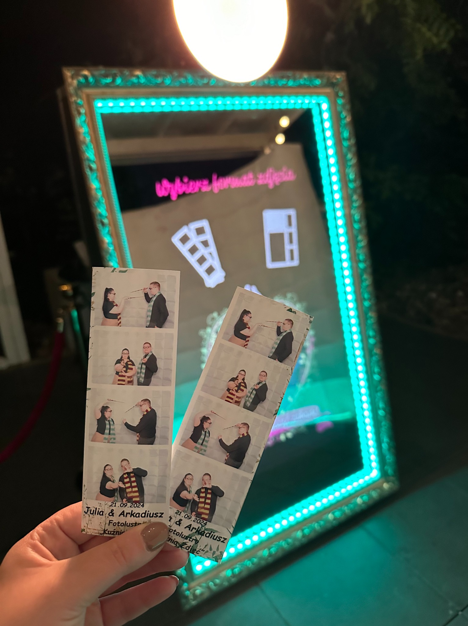Dwie pamiątkowe fotostrip z sesji w fotobudce, trzymane w dłoni na tle lustra z neonowym obramowaniem. Tematyczna sesja pary, klimat Harry'ego Pottera.