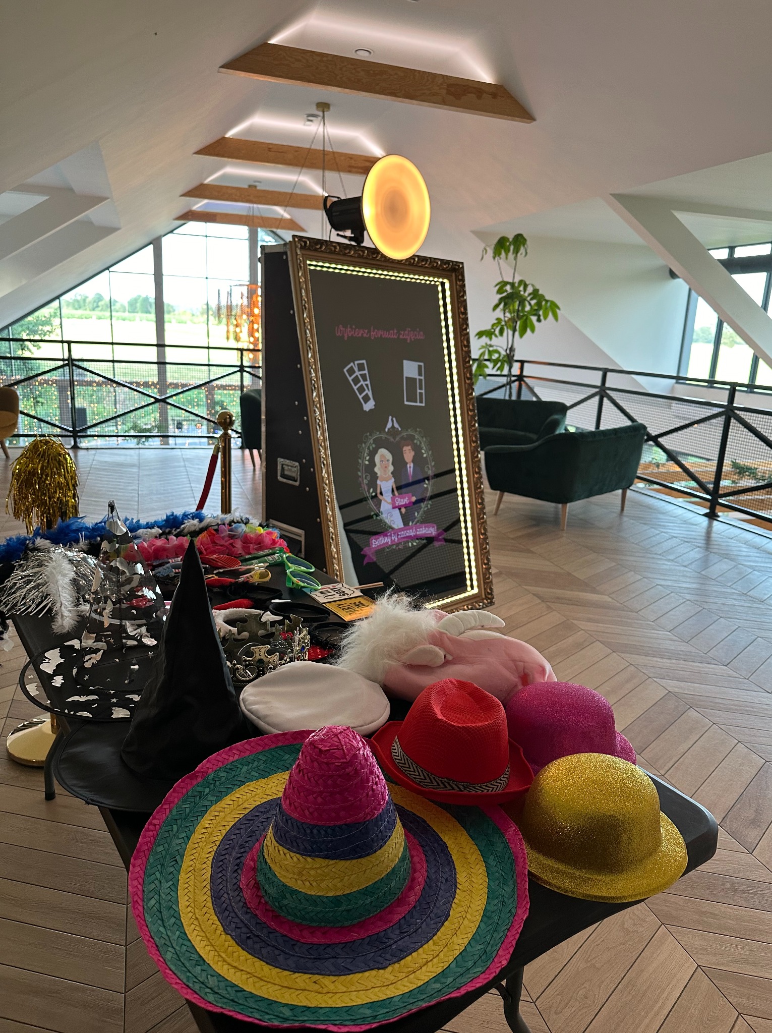 Fotobudka z lustrem i rekwizytami: kolorowe kapelusze, boa, maski na stole. W tle widoczne eleganckie wnętrze z oknami i roślinami.