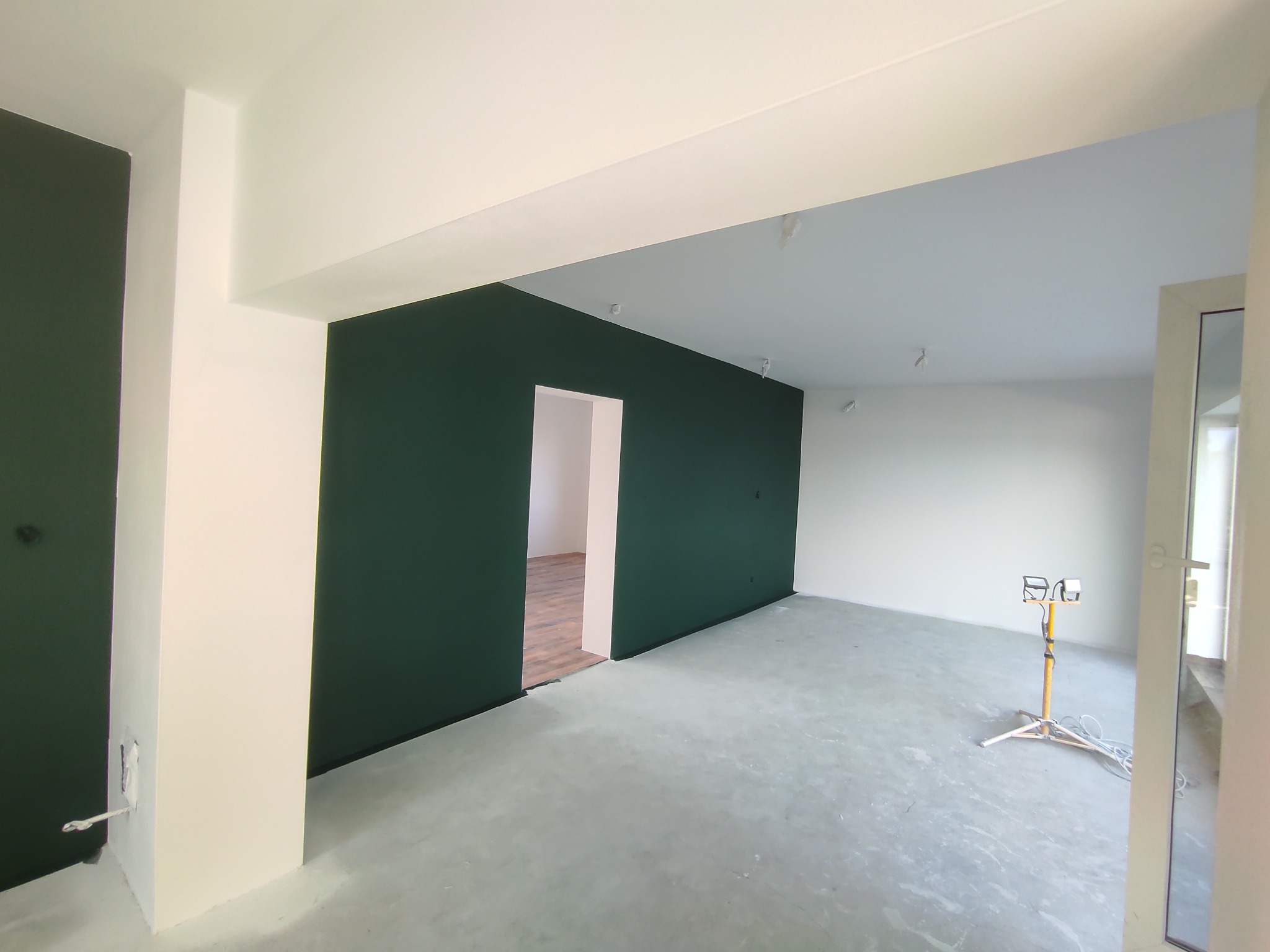 Wnętrze po malowaniu: ściany w odcieniach bieli i butelkowej zieleni, widoczne ślady po szpachlowaniu przy kolumnie. Na podłodze widoczna lampa budowlana.