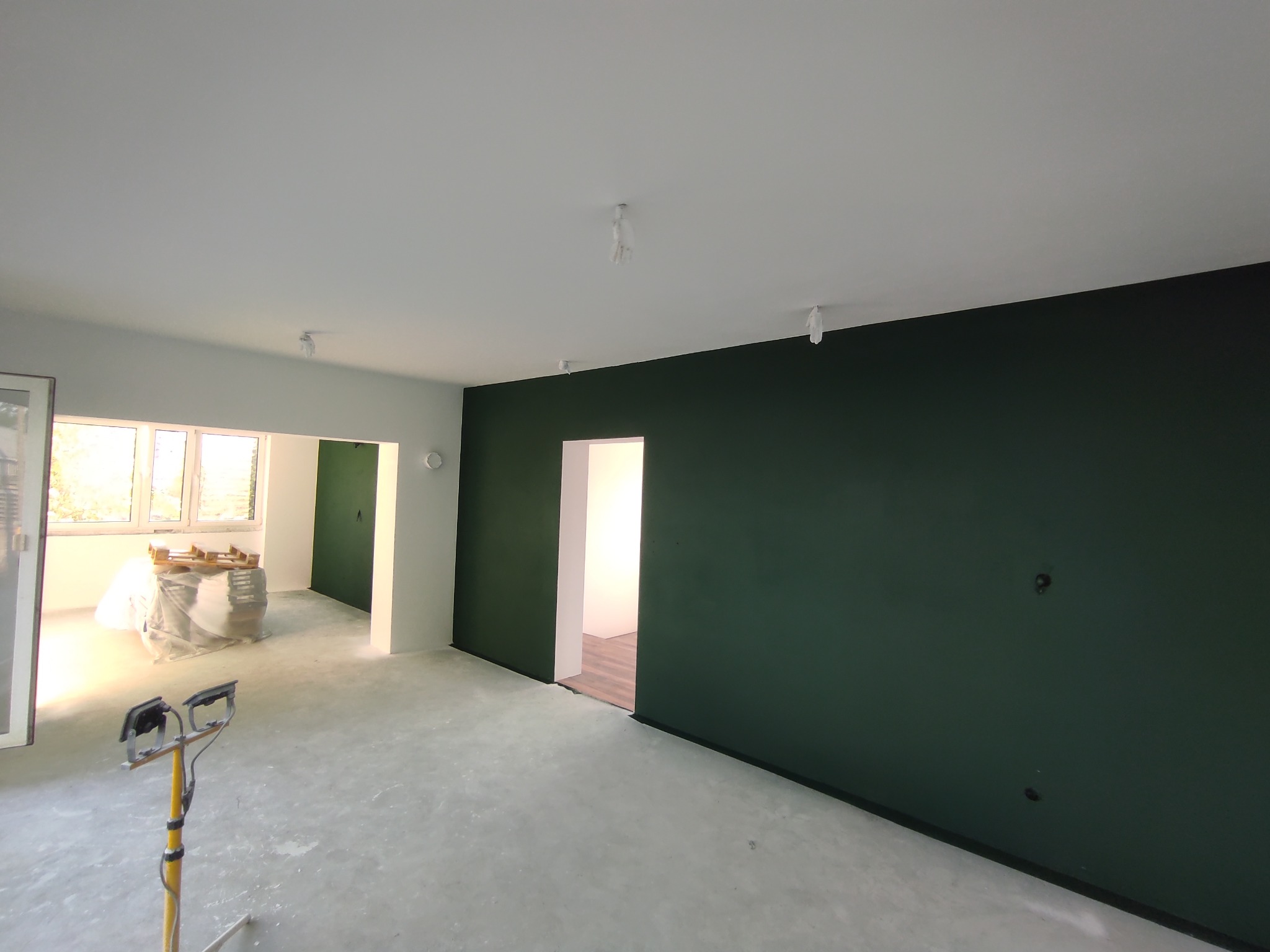 Pomieszczenie po malowaniu: ściana w odcieniach zieleni, zabezpieczone lampy sufitowe, podłoga przygotowana do dalszych prac. Widoczne narzędzia malarskie.